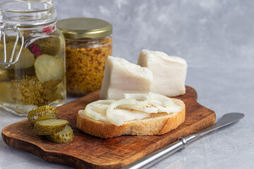 Pickled cucumbers in a glass jar on a wooden board and a sandwich with bacon. Mustard beans in the background. Fermented food.