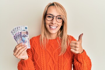 Beautiful blonde woman holding russian ruble banknotes smiling happy and positive, thumb up doing excellent and approval sign