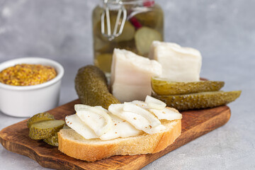 Pickled cucumbers in a glass jar on a wooden board and a sandwich with bacon. Mustard beans in the background. Fermented food.