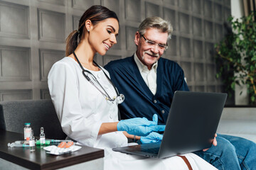 A smiling nurse visits an older man's home, she points a finger at the computer about the test results. Doctor's visit home, remote treatment at home