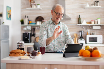 Mature man waving while having a conversation during video call in kitchen enjoying breakfast....