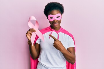 Young african american girl wearing superhero costume holding pink cancer ribbon smiling happy...