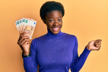 Young african american girl holding bunch of 50 euro banknotes screaming proud, celebrating victory and success very excited with raised arm