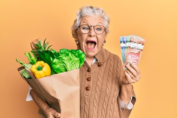 Senior grey-haired woman holding groceries and canadian dollars celebrating crazy and amazed for...