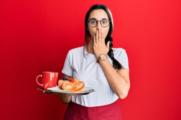 Young hispanic woman wearing waitress apron holding tray with breakfast covering mouth with hand, shocked and afraid for mistake. surprised expression