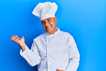 Mature middle east man wearing professional cook uniform and hat smiling cheerful presenting and pointing with palm of hand looking at the camera.