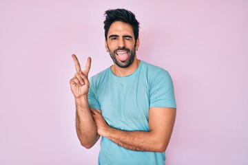 Handsome hispanic man with beard wearing casual clothes smiling with happy face winking at the camera doing victory sign. number two.