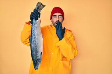 Handsome hispanic man with beard wearing fisherman equipment covering mouth with hand, shocked and...