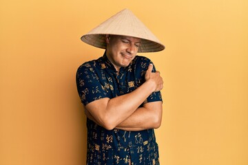 Middle age bald man wearing traditional asian straw hat hugging oneself happy and positive, smiling confident. self love and self care