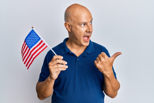 Middle age bald man holding united states flag pointing thumb up to the side smiling happy with open mouth