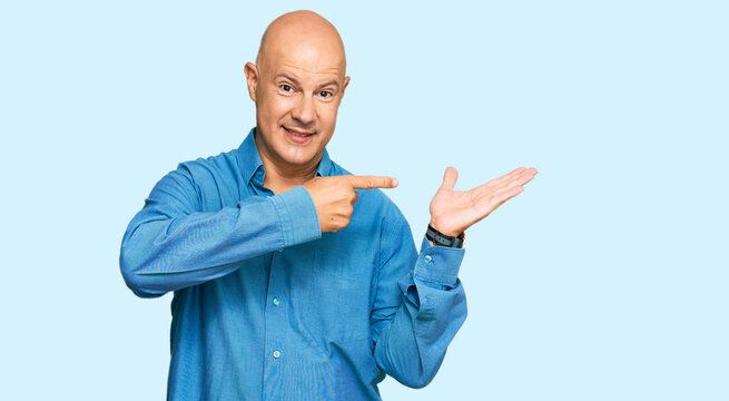 Middle age bald man wearing casual clothes amazed and smiling to the camera while presenting with hand and pointing with finger.