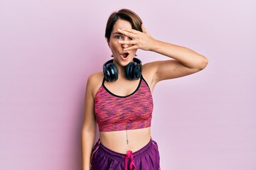 Young brunette woman with short hair wearing sportswear and headphones peeking in shock covering face and eyes with hand, looking through fingers with embarrassed expression.