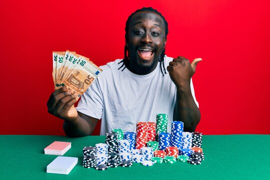 Handsome young black man playing poker holding 50 euros banknotes pointing thumb up to the side smiling happy with open mouth