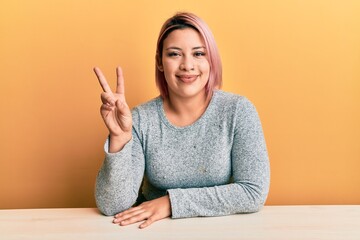 Hispanic woman with pink hair wearing casual clothes sitting on the table smiling with happy face...