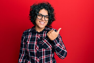 Young hispanic woman with curly hair wearing casual clothes and glasses smiling with happy face looking and pointing to the side with thumb up.