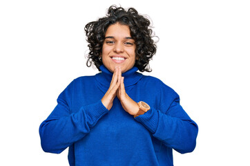 Young hispanic woman with curly hair wearing turtleneck sweater praying with hands together asking for forgiveness smiling confident.