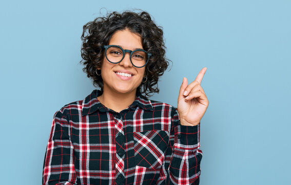 Young hispanic woman with curly hair wearing casual clothes and glasses smiling happy pointing with hand and finger to the side