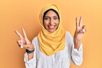 Young brunette arab woman wearing traditional islamic hijab scarf smiling with tongue out showing fingers of both hands doing victory sign. number two.