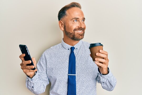 Handsome middle age business man using smartphone and drinking a cup of coffee smiling looking to the side and staring away thinking.