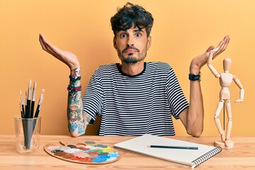 Young hispanic man sitting on the table at art studio clueless and confused expression with arms...