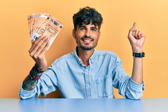 Young hispanic man holding 50 euro banknotes sitting on the table smiling happy pointing with hand and finger to the side
