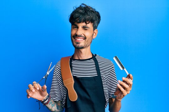 Young hispanic man wearing barber apron holding razor and scissors winking looking at the camera with sexy expression, cheerful and happy face.