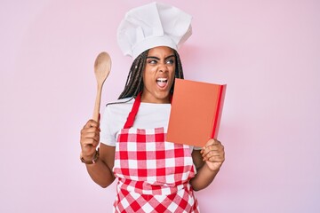 Young african american woman with braids wearing professional baker apron reading cooking recipe...
