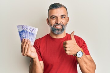 Middle age handsome man holding 20 swedish krona banknotes smiling happy and positive, thumb up doing excellent and approval sign