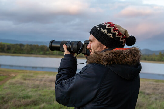 Photo of a attractive and young photographer in the middle of the wild looking through his camera zoom lens. Wearing winter clothes. Explorer, looking for animals