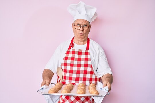 Senior grey-haired man wearing baker uniform holding homemade bread making fish face with mouth and squinting eyes, crazy and comical.