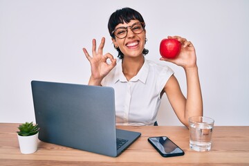 Beautiful brunettte woman working at the office eating healthy apple doing ok sign with fingers,...