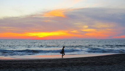 Puerto Escondito, Mexico - December 28 2018 - A woman walking down the beach in front of a beautiful red and orange sunset over the Pacific Ocean in Puerto Escondito, Mexico.  Image has copy space.