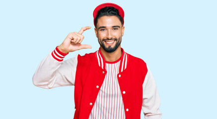 Young man with beard wearing baseball uniform smiling and confident gesturing with hand doing small size sign with fingers looking and the camera. measure concept.