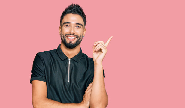Young man with beard wearing sportswear with a big smile on face, pointing with hand and finger to the side looking at the camera.
