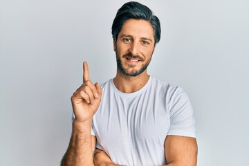 Young hispanic man wearing casual white t shirt smiling with an idea or question pointing finger up with happy face, number one