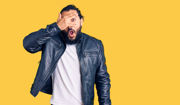 Young arab man wearing casual leather jacket peeking in shock covering face and eyes with hand, looking through fingers with embarrassed expression.