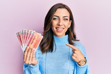 Young brunette woman holding norwegian krone banknotes smiling happy pointing with hand and finger