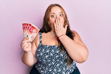 Plus size caucasian young woman holding 20 israel shekels banknotes covering mouth with hand,...
