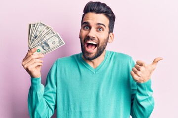 Young handsome man with beard holding bunch of dollar pointing thumb up to the side smiling happy with open mouth