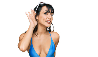 Naklejka premium Young hispanic woman wearing swimsuit and sunglasses smiling with hand over ear listening an hearing to rumor or gossip. deafness concept.
