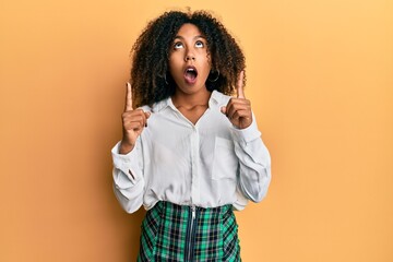 Beautiful african american woman with afro hair wearing scholar skirt amazed and surprised looking up and pointing with fingers and raised arms. - obrazy, fototapety, plakaty