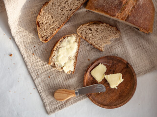 Homemade fresh wheat-rye bread on sourdough on a natural color linen napkin with butter in a wooden plate with a butter knife on a white background in the morning light