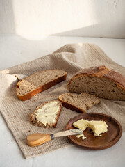 Homemade fresh wheat-rye bread on sourdough on a natural color linen napkin with butter in a wooden plate with a butter knife on a white background in the morning light