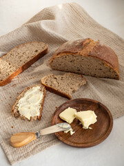 Homemade fresh wheat-rye bread on sourdough on a natural color linen napkin with butter in a wooden plate with a butter knife on a white background in the morning light