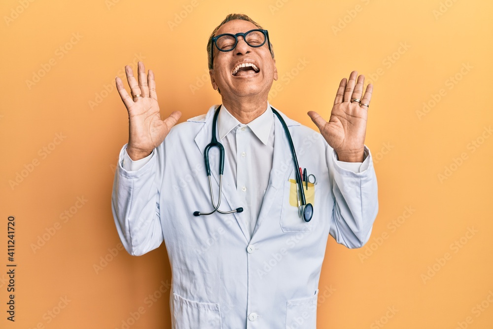 Sticker Middle age indian man wearing doctor coat and stethoscope celebrating mad and crazy for success with arms raised and closed eyes screaming excited. winner concept