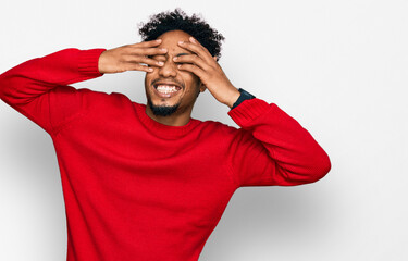 Young african american man with beard wearing casual winter sweater covering eyes with hands smiling cheerful and funny. blind concept.