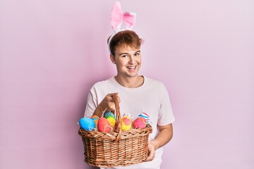 Young caucasian man wearing cute easter bunny ears holding wicker basket with colored eggs looking...