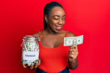 Young african american woman holding jar with savings and 1 dollar banknote winking looking at the...
