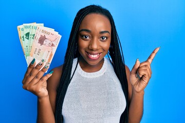 Young african american woman holding south korean won banknotes smiling happy pointing with hand and finger to the side