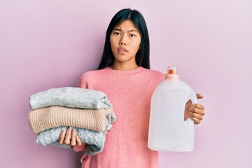 Young chinese woman holding detergent bottle and clothes clueless and confused expression. doubt...